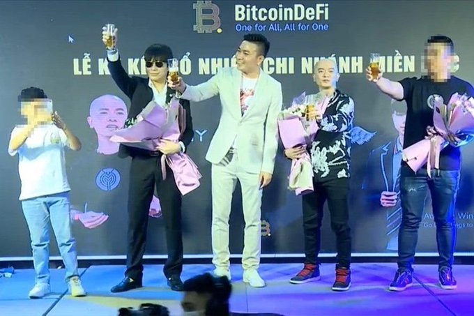 A group of men on stage at a BitcoinDeFi ('One for All, All for One') event, holding champagne glasses and bouquets for a launch celebration, with a Vietnamese banner behind them.