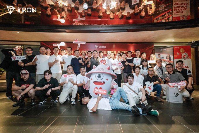 A large group of people poses for a photo at a TRON event, with the TRON logo visible on banners and signs reading "TRX ON TRON" and "TOP Vietnam," along with a pink mascot in the center.