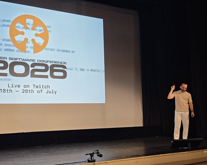 A man stands on a stage in front of a projection screen displaying a logo and text reading '...ER SOFTWARE CONFERENCE 2026, Live on Twitch, 18th - 20th of July'.