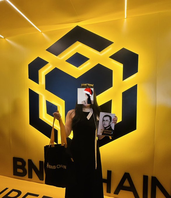 The image features a woman standing in front of a yellow wall with a logo for Bitcoin, which reads "BNCHAIN" at the bottom. She has her face covered by a paper hat or mask, making her appear as if she's hiding from someone or something. The woman is holding a bag and appears to be posing for a photo. There are also two other people in the image who seem to be standing behind the woman.