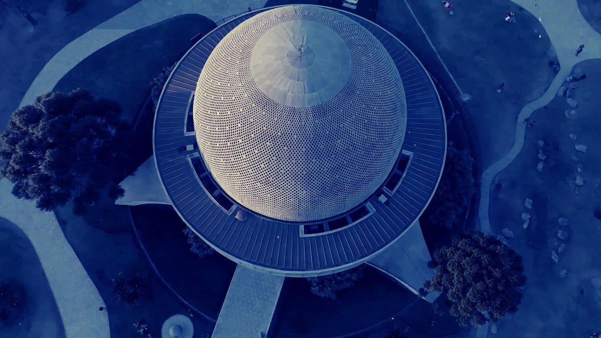 The image features an aerial view of a large dome-shaped building, possibly a museum or a park pavilion with a circular roof. The building appears to be made of concrete and has a unique design that sets it apart from other structures. In the background, there are trees and people walking around, indicating that this location is likely a public space where visitors can enjoy outdoor activities.