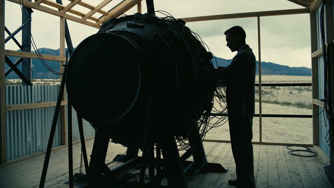 A silhouetted man in a suit stands beside a large spherical mechanical device in an unfinished wooden structure with mountains visible through the windows, suggesting a sci-fi film still.