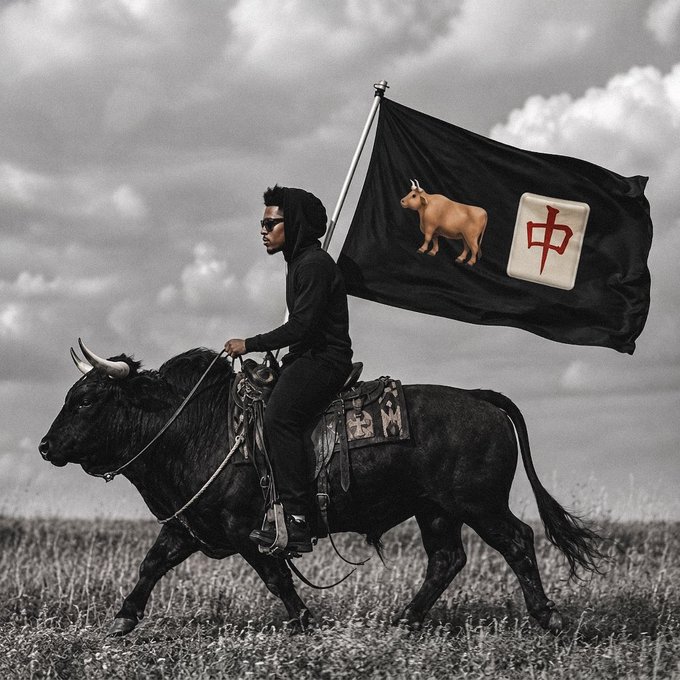 A man in dark clothing and sunglasses rides a black bull across a grassy field, carrying a large flag featuring a cow emoji and the Chinese character "中" (meaning "China"). The image ties into the caption "let's ride. $ANSEM" by depicting a powerful, bold entrance on a bull—symbolizing a strong market move or confident stance toward the $ANSEM token with a nod to its Chinese community.