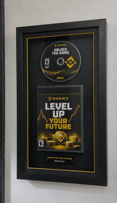 A framed Binance display features "Unlock the Game" on a disc and "Level Up Your Future" on a case cover, with a plaque reading "Building the future of finance since 2017," reinforcing the post's message that Binance is ushering in the next phase of crypto.