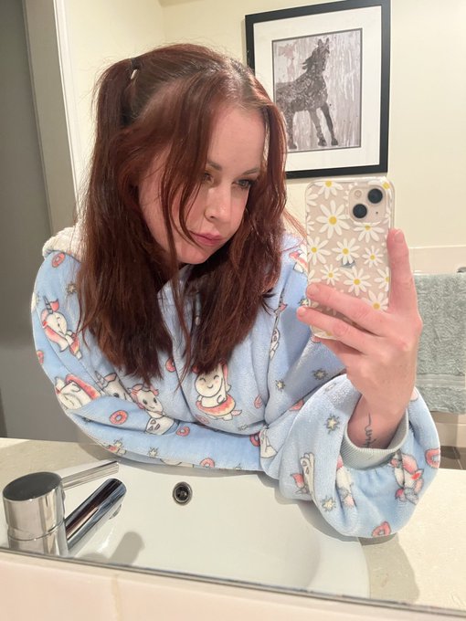 A woman takes a mirror selfie in her bathroom, wearing cozy blue pajamas with a snowman print, her phone case decorated in white daisies. With tousled auburn hair and a soft, contemplative expression, she pairs the candid snapshot with a relatable post about how her good morning took a turn for the worse, leaning into the lesson that "shit happens" and reminding herself (and followers) not to let a bad mood knock her down.