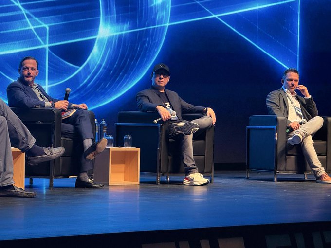 Several panelists sit in black chairs on a stage in front of a blue digital backdrop with a globe graphic; microphones, water bottles, and small wooden side tables are arranged around them.