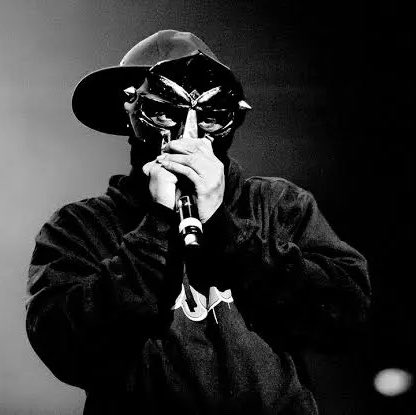 A black and white photo shows a person in a wide-brimmed hat and dark clothing holding a microphone close to their face, with their hand partially obscuring their features.