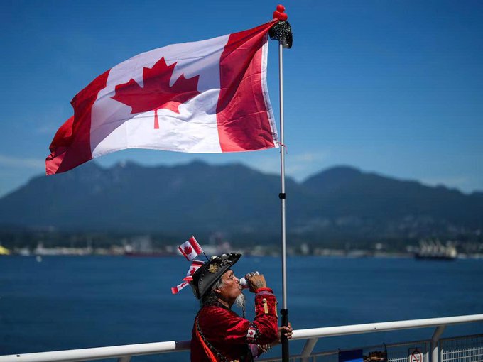 A person wearing a hat adorned with small Canadian flags holds a large Canadian flag on a pole against a backdrop of mountains and water, drinking from a can. The image symbolizes Canadian national identity, which ties to the post about rising cryptocurrency ownership among Canadians, now at 25% according to a recent Ontario Securities Commission survey.