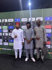 Three men pose together in front of a sponsor backdrop at the NFT__NG event, with one wearing a red cap and white traditional attire, another in a dark outfit, and the third in a dark agbada holding a briefcase, all standing before a wall displaying various brand logos.