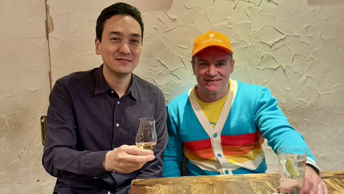 Two men smile at a table during what appears to be a celebratory dinner, one holding a glass of white wine and the other wearing an orange Bitcoin cap alongside a colorful striped cardigan, visually personifying the buyer and seller who struck the ¥11-per-share deal for 70% of a Tokyo-listed company on 16 March 2022.