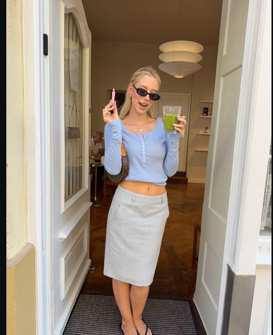 A blonde woman wearing sunglasses, a light blue cardigan, and a striped skirt holds up a pink smartphone and a green drink while standing in a doorway.