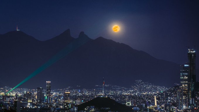 A nighttime cityscape featuring a full orange moon rising over silhouetted mountains, with city lights and a green laser beam visible.