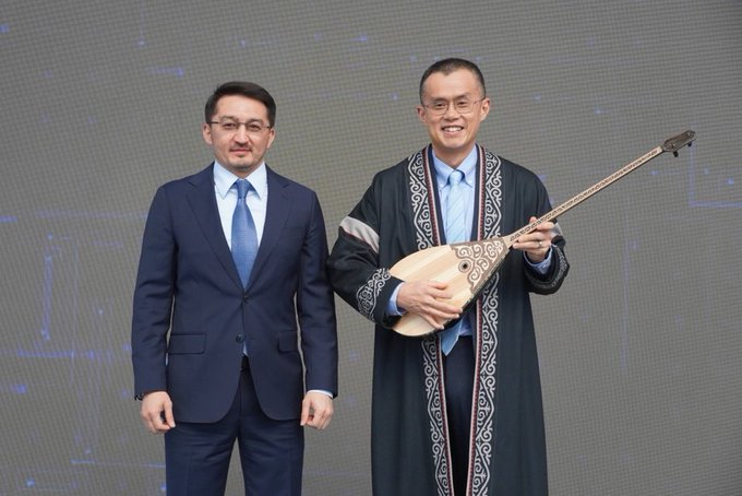 Two men stand together at an event, with the man on the right wearing traditional dark embroidered attire and proudly holding a dombra—a traditional Kazakh stringed instrument—while the man on the left wears a modern navy suit, symbolizing a blend of cultural heritage and contemporary innovation that ties into the post's theme of CZ championing crypto adoption.