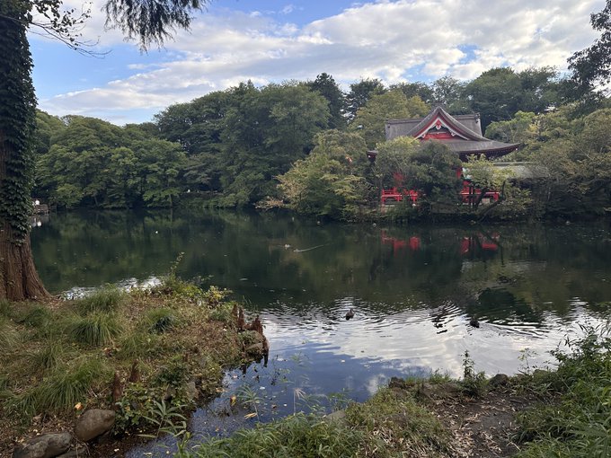 A serene Japanese garden with a red traditional pavilion reflected in a calm pond, surrounded by lush green trees under a partly cloudy sky.