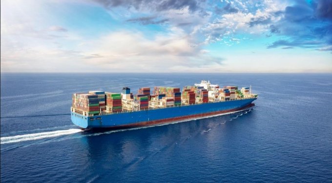 An aerial view of a large blue container ship loaded with multicolored shipping containers sailing through the open ocean under a partly cloudy sky.
