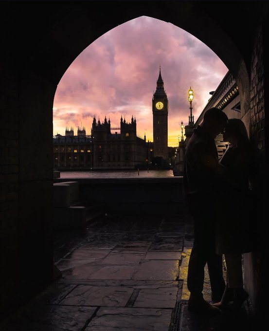 A silhouetted couple embraces under a stone archway, with London's Big Ben and the Houses of Parliament illuminated against a pink and orange sunset sky in the background.