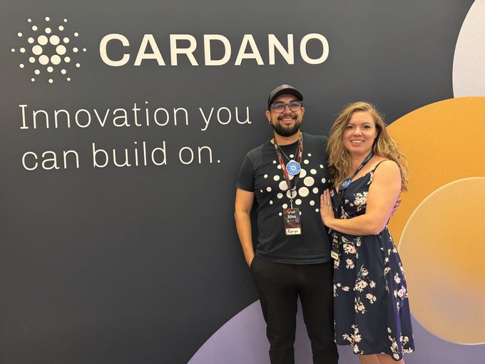 Two people pose in front of a Cardano branded backdrop that reads 'CARDANO - Innovation you can build on.'
