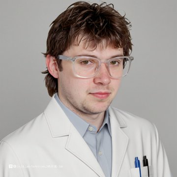 A studio portrait of a young man with brown hair wearing clear-framed glasses, a white lab coat, and a light blue shirt, with pens in his coat pocket.