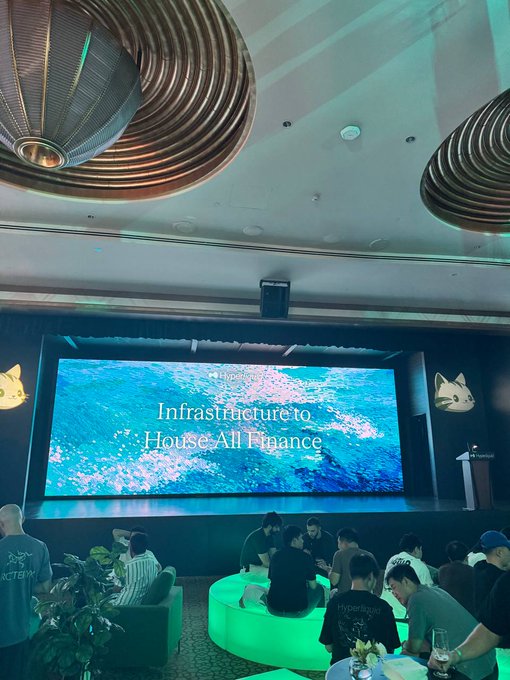 A conference or event space features a large screen displaying 'Hyperliquid: Infrastructure to House All Finance' with cat logos, while attendees sit at a circular glowing table below decorative ceiling fixtures.