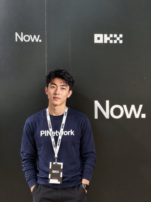 A young man wearing a "PINetwork" sweatshirt and an "OKX" lanyard poses in front of a black wall featuring the "OKX" logo and the word "Now." repeated—visually representing the PINetwork-OKX partnership highlighted in the post about the crypto industry's "casino" landscape and PI's mission.