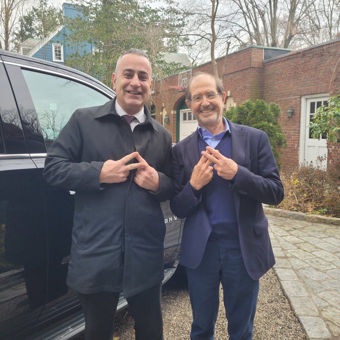The image shows two men standing next to a car, both wearing suits and ties. One man is smiling while the other is making a peace sign with his fingers. The background includes a brick building and trees, creating an outdoor setting for their photo.