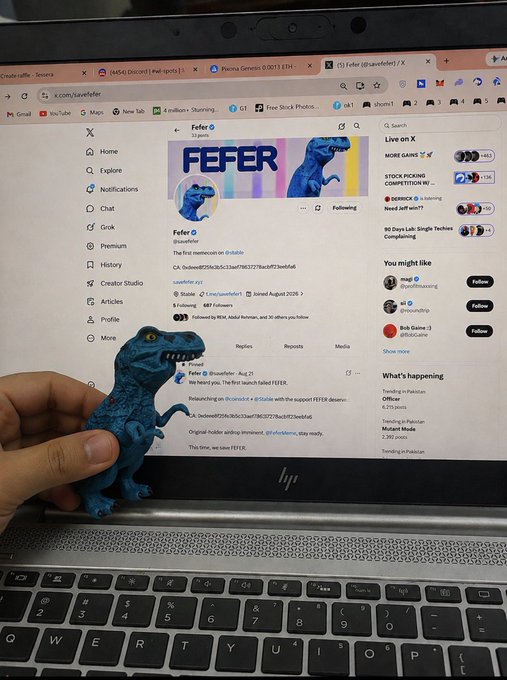 A photograph of a small blue toy dinosaur held up in front of a laptop screen displaying the X (formerly Twitter) profile for the account '@sawfeer', showing their header image, follower count, and pinned post about a meme coin launch.