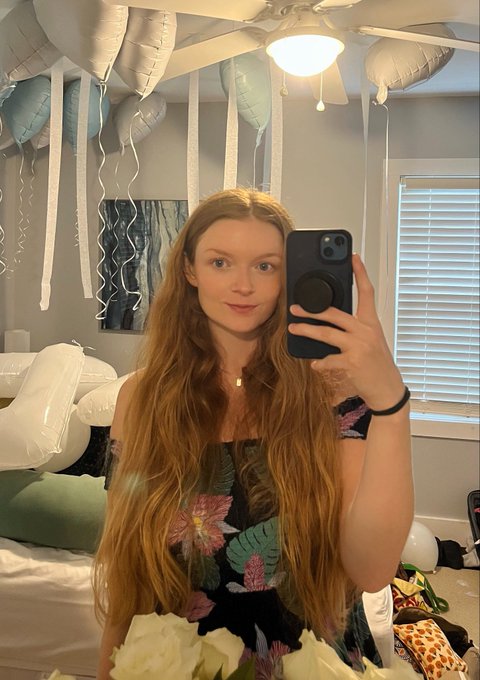 A young woman with long red hair takes a mirror selfie in a room decorated with white balloons and streamers.