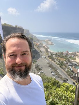 A bearded man in a white t-shirt takes a selfie with a coastal cliffside road, beach, and ocean visible in the background.