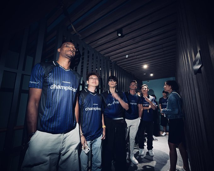A group of players wearing matching blue "champions" jerseys gather in a dimly lit hallway, with one holding a microphone as they prepare for the $150,000 competition hosted by Coinbase.