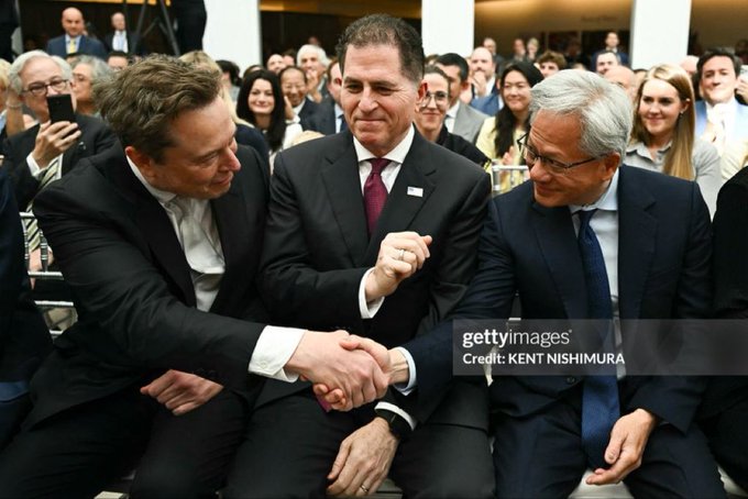 A Getty Images photo by Kent Nishimura showing Elon Musk shaking hands with another man while a third man looks on, with an audience in the background.