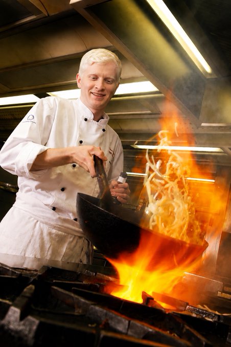 A white-haired chef in a traditional white uniform smiling while flambéing food in a wok over an open gas flame in a commercial kitchen.