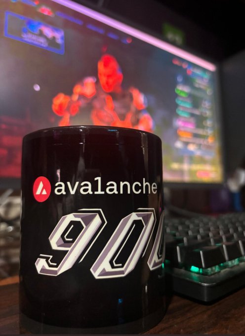 A black mug with 'avalanche' branding and the number '900' in large white text sits on a desk in front of a mechanical keyboard and a computer monitor displaying a colorful game or interface.