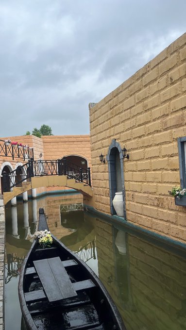A black gondola boat floats in a narrow canal beside a tan brick building with arched windows and a decorative bridge, under an overcast sky.