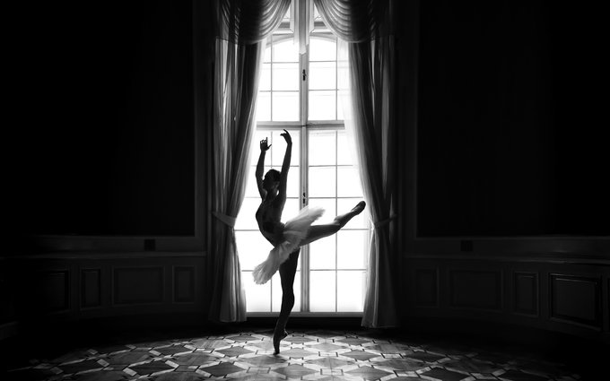 A black and white photograph captures the silhouette of a ballerina in a tutu performing an elegant pose on pointe in front of a large, bright window with sheer curtains.