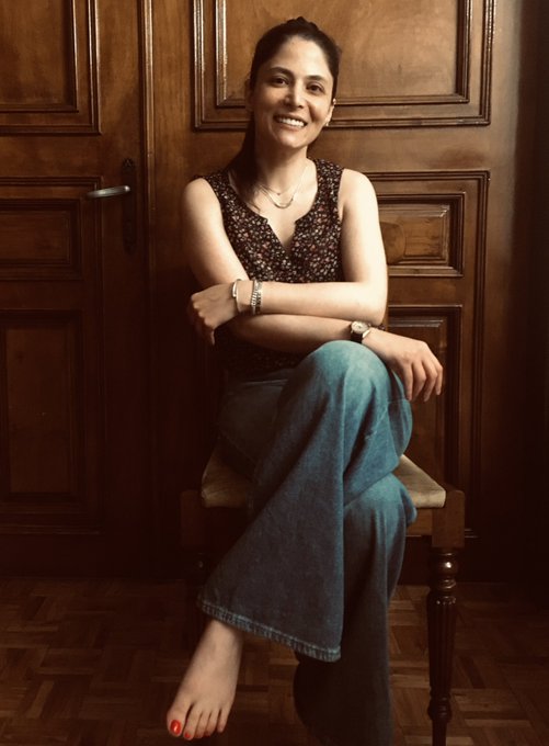 A smiling young woman with dark hair sitting barefoot on a wooden chair, wearing a dark floral top and loose blue jeans, positioned in front of an ornate wooden door.