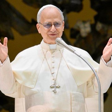 Pope Leo XIV in white papal vestments and glasses, wearing a cross necklace, speaking at a microphone with both hands raised in a welcoming gesture.