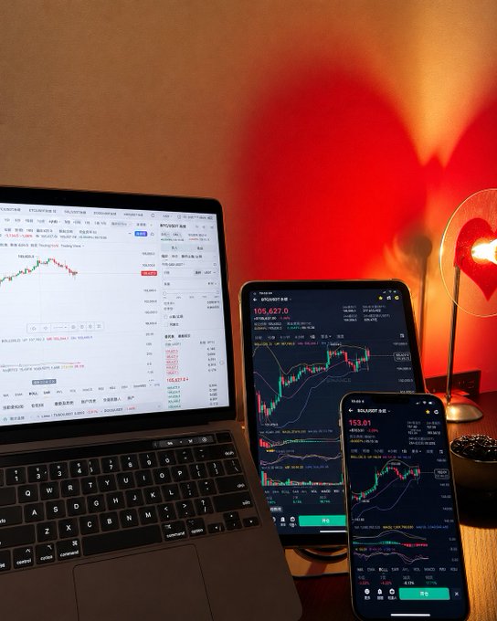 The image shows a laptop screen displaying stock price graphs for Bitcoin and Ethereum. Beside the laptop are three mobile phones, each displaying different stock market analysis results. The scene takes place on a desk with a lamp casting an eerie red light over the monitors.