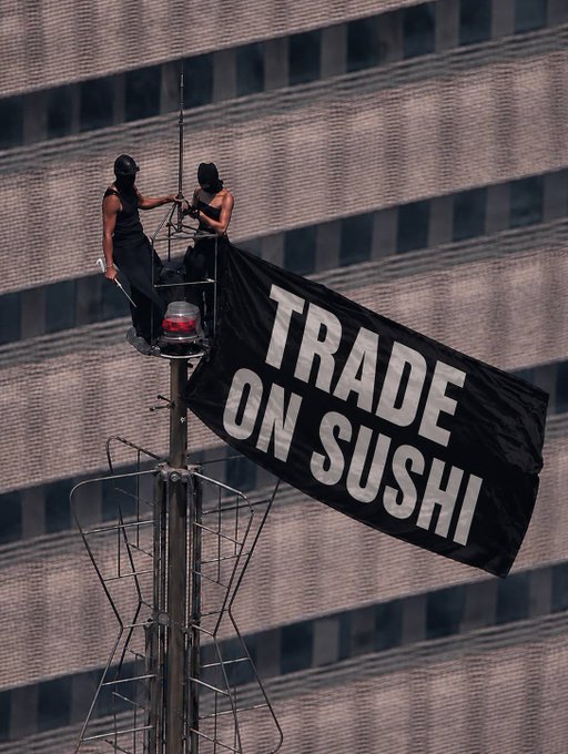Two masked activists perch atop a tall antenna near a high-rise building, unfurling a large black banner reading **"TRADE ON SUSHI"** as they appear to contemplate their precarious climb back down.

The image humorously illustrates the post's caption "Ok, how do I get down now?"—capturing the very real dilemma of activists who climbed to a dangerous height for a publicity stunt, only to face the daunting descent back to the ground.