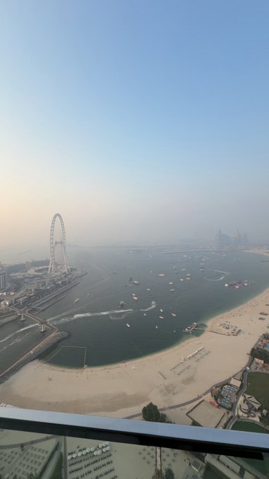 Aerial view from a high-rise balcony overlooking a beach with a large Ferris wheel, boats in the water, and hazy coastal cityscape.