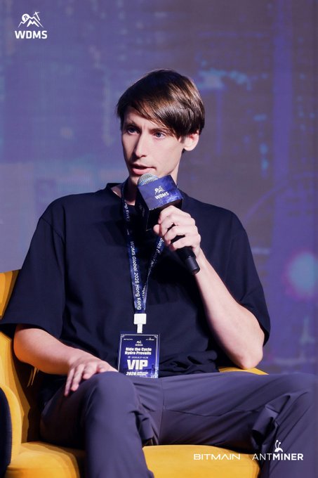 A young man wearing a "VIP" badge speaks into a microphone at a WDMS (World Digital Mining Summit) event sponsored by Bitmain AntMiner, with his badge identifying him as from the "Make the Crypto" panel.