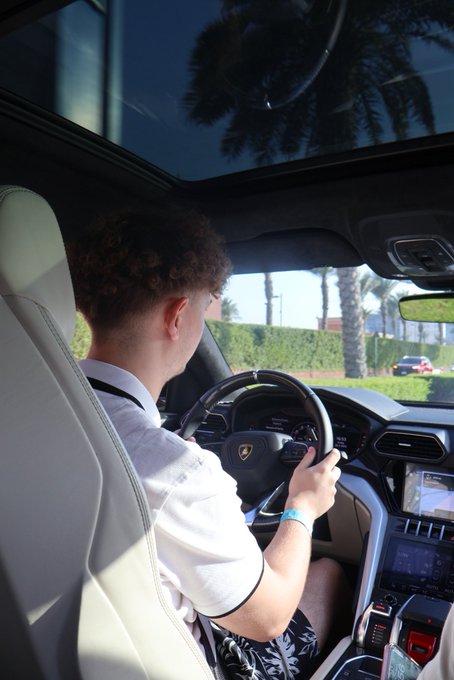 A curly-haired man in a white polo shirt drives a Lamborghini, with the iconic bull logo visible on the steering wheel. The post "GM if you GM" pairs with the image as a casual greeting inviting others to reply with their own "good morning" while showcasing a luxury lifestyle.