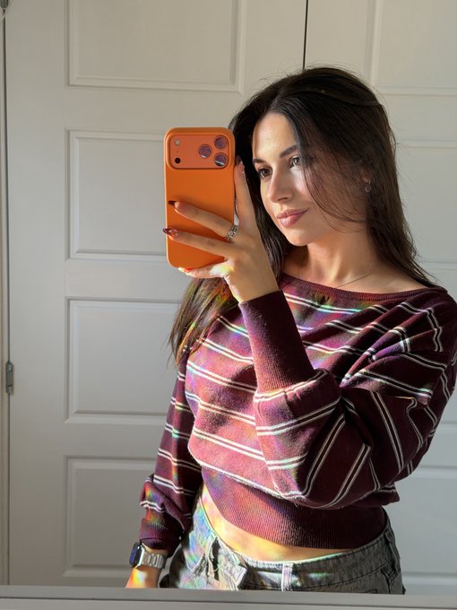 A young woman with long brown hair takes a mirror selfie holding an orange iPhone, wearing a purple striped cropped sweater and jeans, as she celebrates "Happy @RuneFest Day!" in casual, festive style.