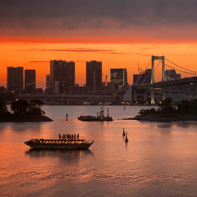 A scenic sunset view over Tokyo Bay featuring the Rainbow Bridge, silhouetted city skyline, and boats on calm waters under an orange sky.