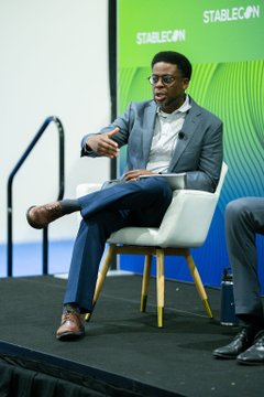 A man in a gray blazer and dark pants speaks while seated on a white chair on a stage at the 'STABLECON' conference.