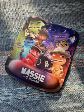 The image features a colorful mouse pad with cartoon characters, including a character named "Wassie" who appears to be a monster or a character from a video game. The mouse pad has been placed on top of a gray rug, which is spread out in front of it. There are also some text elements present in the image that read "looking for smol loot? We're cracking open the fridge for a physical + digital wassie loot drop". This suggests that there might be a game or an event related to video games taking place, and the mouse pad could potentially serve as a prize or a collectible item.