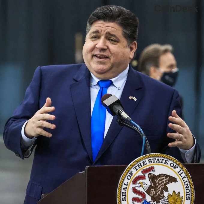 The image shows a man wearing a suit and tie standing at a podium with his hands outstretched as if he's giving a speech or addressing an audience. There are two people visible behind him, one of whom appears to be wearing a mask. The background features a large logo that reads "SEAL OF THE STATE OF THE STATE OF VIOLA".