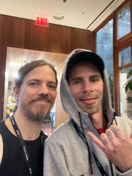 Two men take a selfie indoors, with the man on the right wearing a gray hoodie and baseball cap and flashing a hand gesture with visible finger tattoos, while both wear lanyards—one reading "HIMSS24"—matching the post's optimistic "GM" greeting vibe.