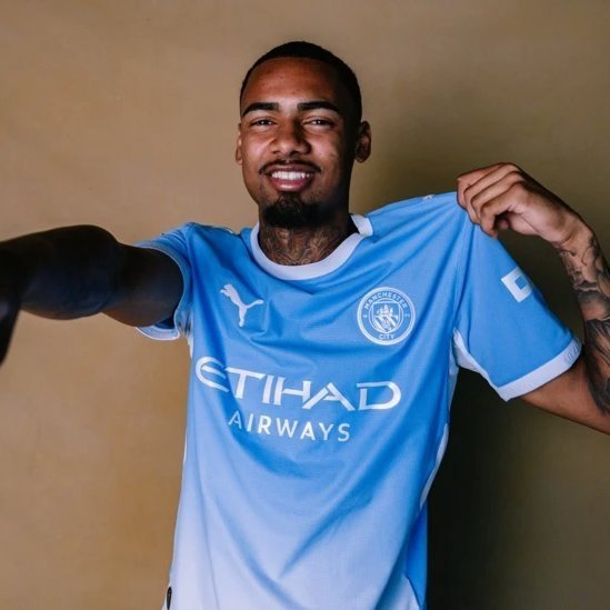 Football player Gabriel Jesus smiling and holding up a light blue Manchester City home jersey featuring the Etihad Airways sponsor and Puma logo.