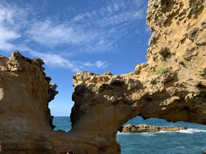 A coastal landscape featuring dramatic, weathered limestone rock formations with a natural arch, set against a blue sky and ocean with waves.