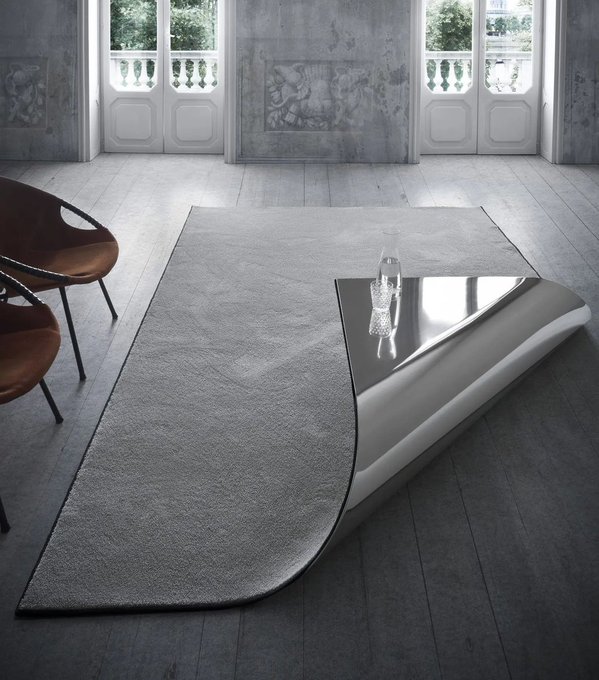 A modern living room features a large gray area rug with a uniquely shaped black and white coffee table on top, accompanied by brown leather chairs and tall windows in the background.